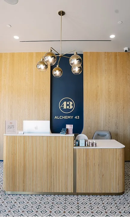 Alchemy 43 short-form post: a wood-slat reception desk with brass pendant lights and a navy logo wall in a bright clinic.