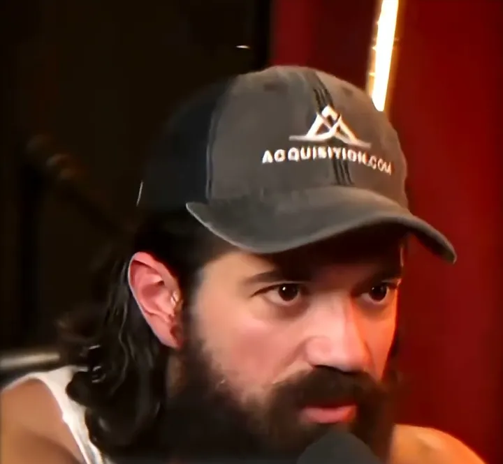 Alex Hormozi short-form post: a bearded man in an Acquisition.com cap in front of a podcast microphone and a red studio curtain.