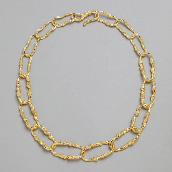 Alexis Bittar short-form post: a gold Brut Link chain necklace with textured oval links laid in a ring on a grey background.