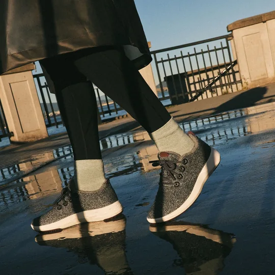 Allbirds short-form post: a person walking in dark Wool Runner Mizzles sneakers over a wet pavement beside a railing in low sun.