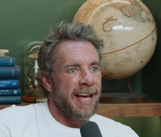 Armchair Expert short-form post: a bearded man in a white t-shirt talking at a microphone in a green-walled room with a globe.