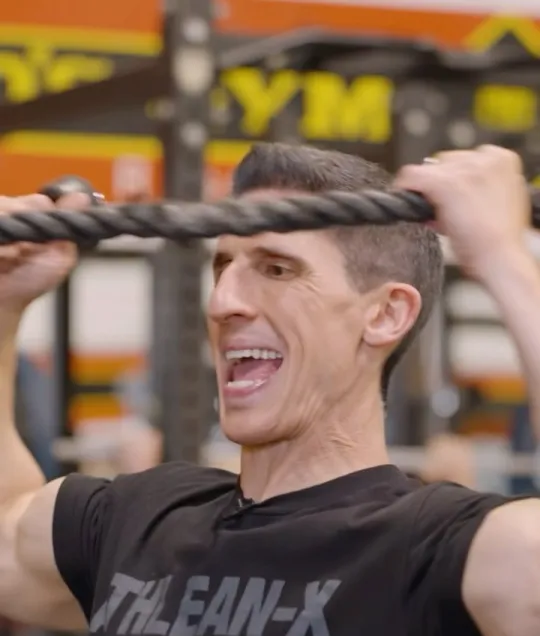 ATHLEAN-X short-form post: a man in a black ATHLEAN-X shirt pulling a rope toward his face in a gym.