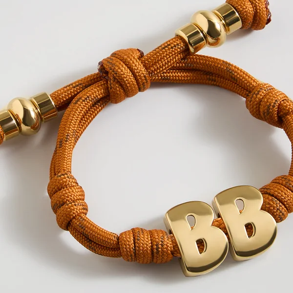 BaubleBar short-form post: a toffee-orange knotted cord bracelet with two gold bubble-letter B beads on a light grey background.
