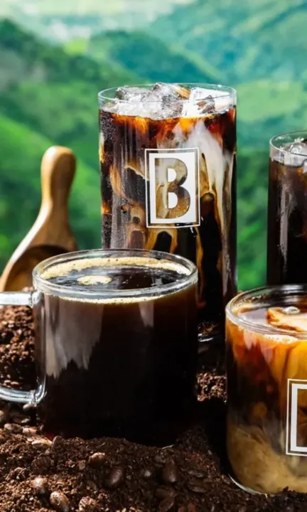 BIGGBY Coffee short-form post: three glasses of iced coffee with ice and cream set on dark soil in a green forest.