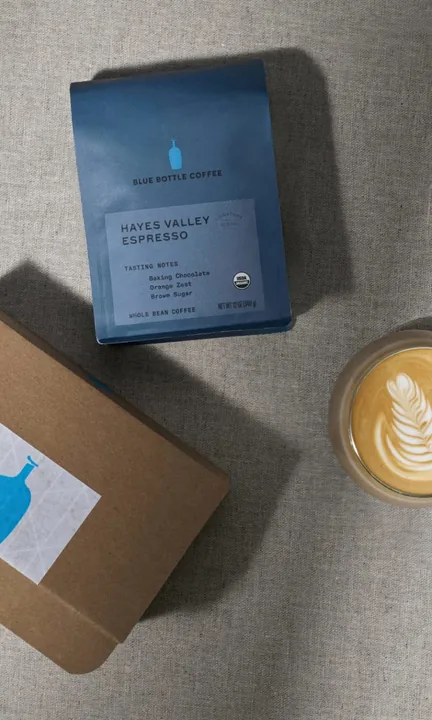 Blue Bottle Coffee short-form post: a blue coffee bag and a kraft box beside a latte with latte art on a stone surface.