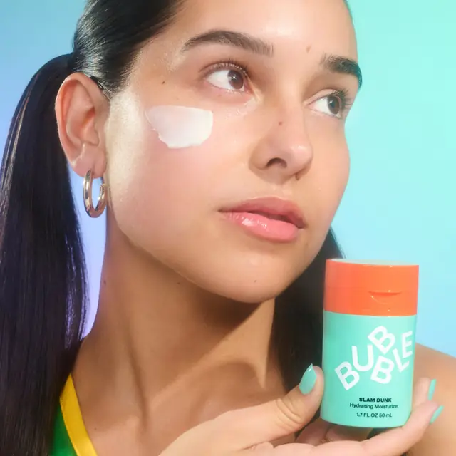 Bubble short-form post: a woman with moisturizer on her cheek holding the teal and orange Slam Dunk jar against a bright blue background.