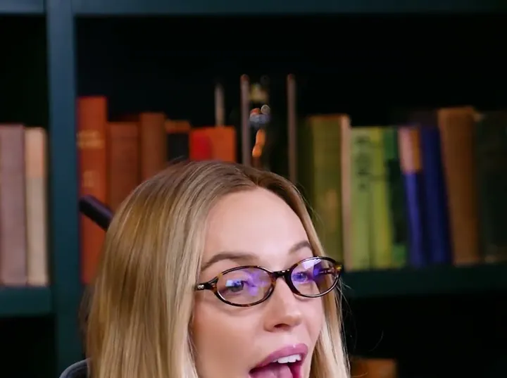 Call Her Daddy short-form post: a woman with long blonde hair and glasses in a blue sweatshirt speaking at a Call Her Daddy microphone.