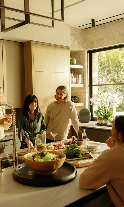 Coldwell Banker short-form post: four friends cooking and talking around a kitchen island beside a large window.