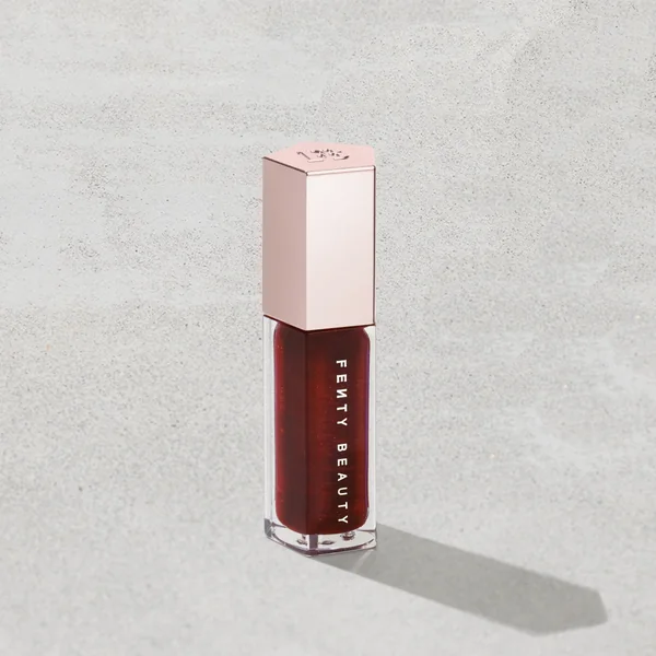 Fenty Beauty short-form post: a Gloss Bomb lip luminizer in a deep berry shade with a pink cap, standing on a pale grey stone surface.