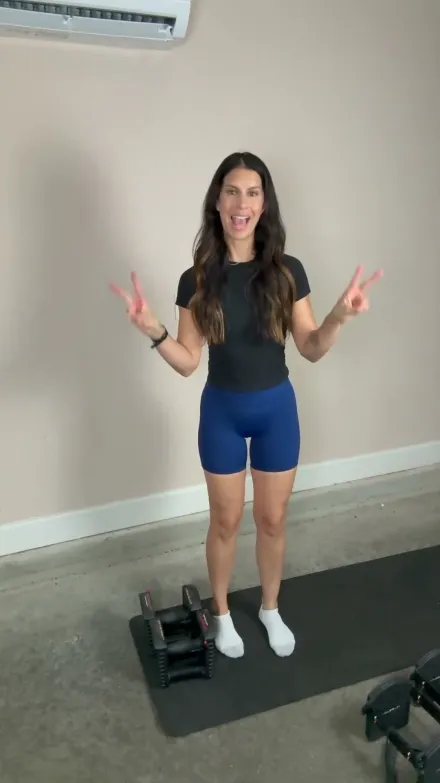 Fitness Blender short-form post: a smiling woman in a black top and blue shorts waving next to dumbbells on a mat.
