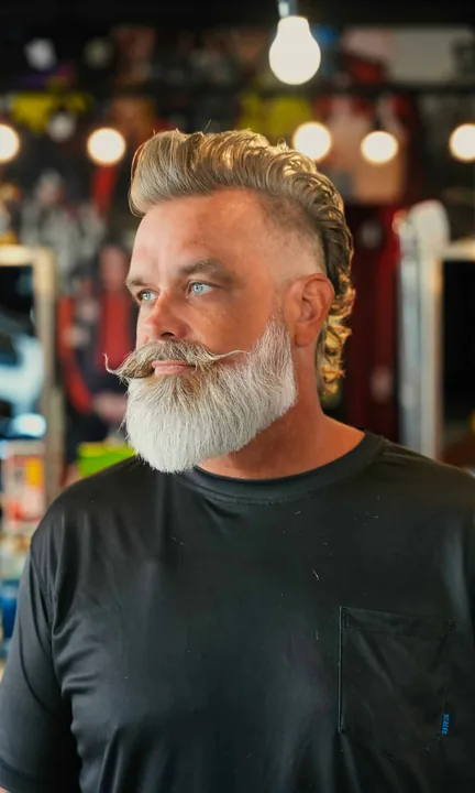 Floyd's 99 Barbershop short-form post: a man with a white beard and styled hair in a barbershop with warm bulbs behind him.