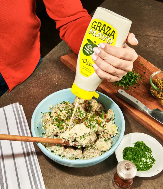 Graza short-form post: a hand squeezing Original Mayo from a yellow Graza bottle over a bowl of potato salad.