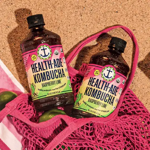 Health-Ade short-form post: two Raspberry Lime kombucha bottles resting in a pink mesh bag on sand with limes.