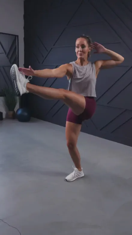Heather Robertson short-form post: a woman in a grey top and plum shorts doing a standing leg raise in a dark studio.