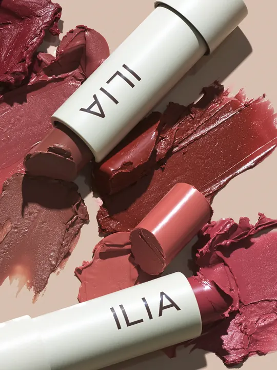 ILIA short-form post: a flat lay of white Balmy Tint lip balm sticks and smeared rosy-brown color on a blush-pink surface.