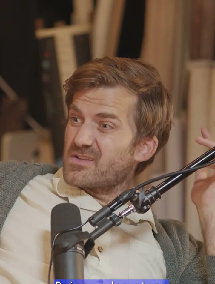 Johnny Harris short-form post: a man in a beige jacket speaking at a podcast microphone.