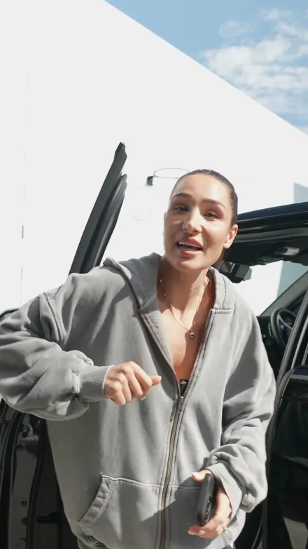 Kayla Itsines short-form post: a woman in a grey zip hoodie talking to camera in front of a car and a bright wall.