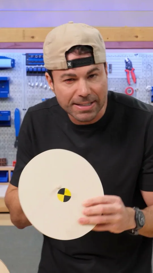 Mark Rober short-form post: a man in a backwards cap holding a round board with a crash-test target on it in a workshop.