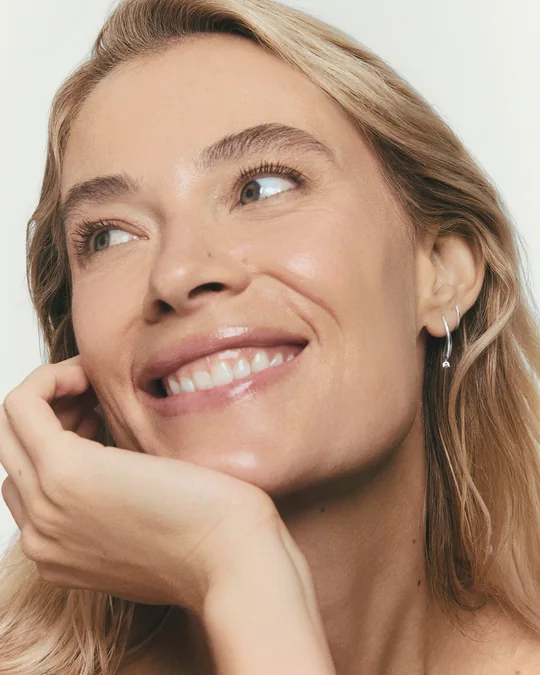 Mejuri short-form post: a smiling woman resting her chin on her hand, wearing a silver Open Dome twist earring.