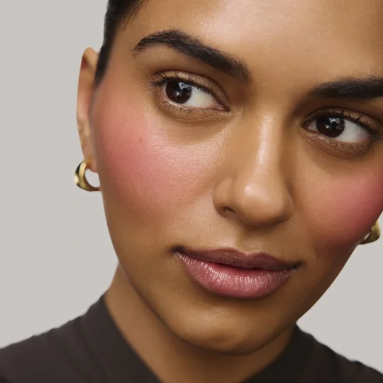 Merit short-form post: a close-up of a woman glancing to the side with a soft pink flush of balm on her cheek.