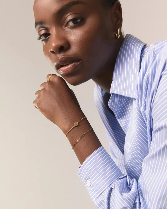 Missoma short-form post: a woman in a blue striped shirt resting her chin on her fist, wearing slim gold bracelets.
