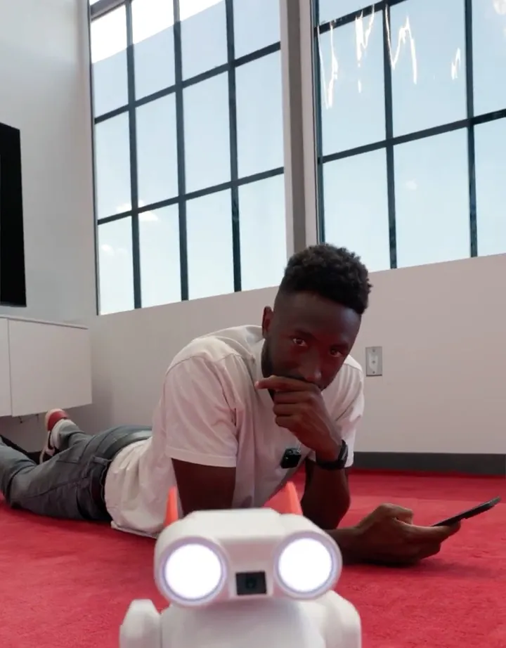 Marques Brownlee short-form post: a man lying on a red floor next to a small white robot with glowing eyes.