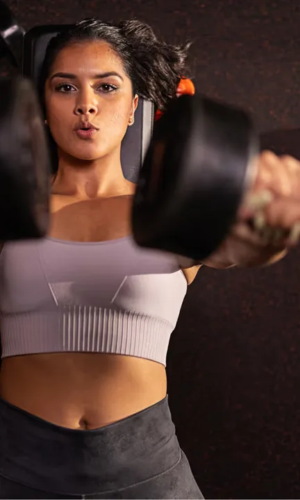 Orangetheory Fitness short-form post: a woman pressing a pair of dumbbells above her chest against a dark background.