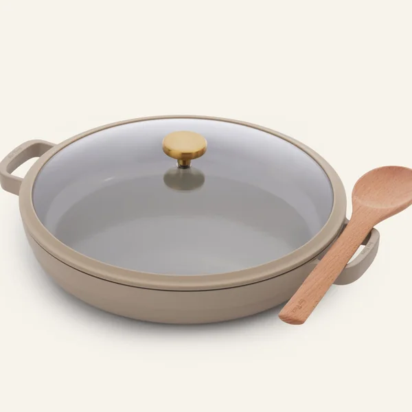 Our Place short-form post: a warm taupe ceramic nonstick Always Pan with a glass lid, a gold knob and a wooden spoon on a cream background.