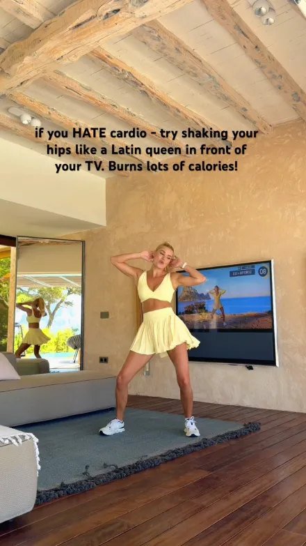 Pamela Reif short-form post: a woman in a yellow outfit dancing on a mat in front of a TV in a bright room.
