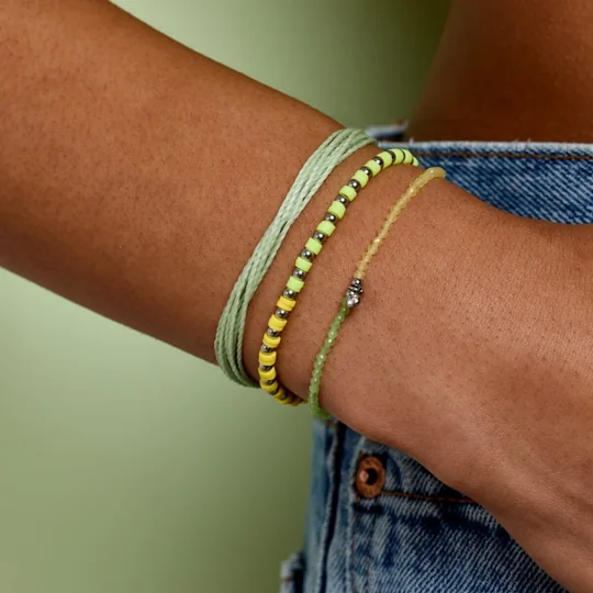 Pura Vida short-form post: a wrist wearing a stack of mint green and yellow friendship bracelets against a green backdrop and denim.