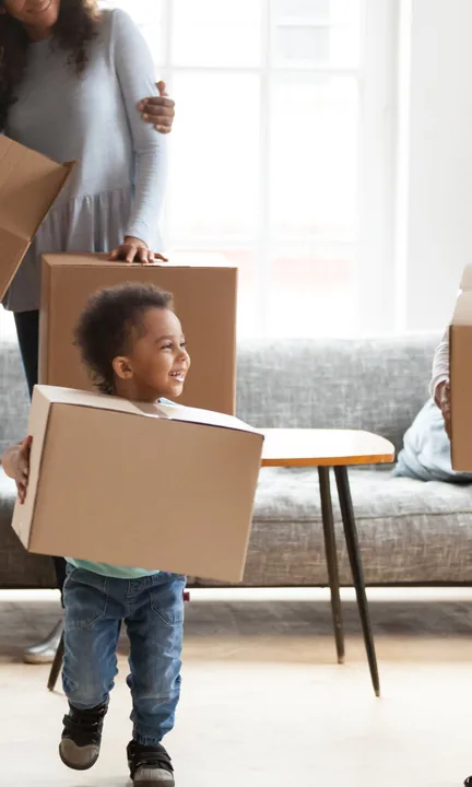 RE/MAX short-form post: a toddler carrying a moving box through a bright living room as an adult carries another behind him.