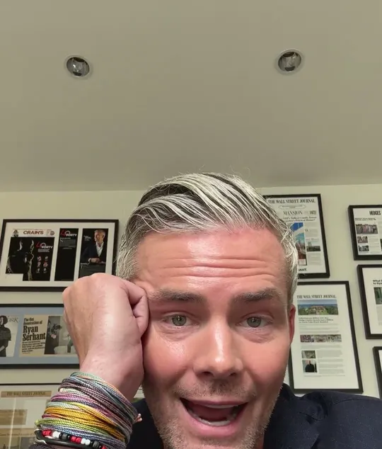 Ryan Serhant short-form post: a grey-haired man in a dark jacket smiling with a hand on his cheek, in an office with framed photos on the wall.