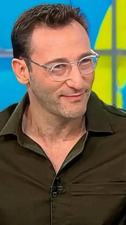 Simon Sinek short-form post: a man in an olive shirt and clear glasses sitting on a TV studio set, smiling slightly.